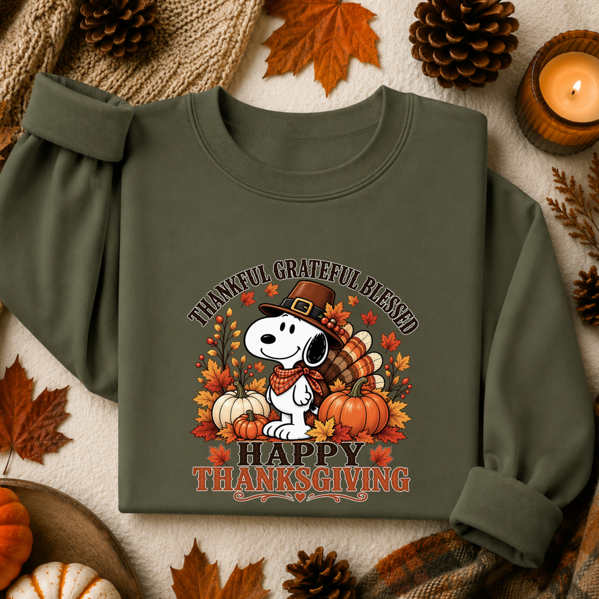 An olive green long-sleeve sweatshirt featuring a festive Thanksgiving graphic of Snoopy with pumpkins, autumn leaves, and the text 'THANKFUL GRATEFUL BLESSED HAPPY THANKSGIVING'. The sweatshirt is styled in a flat-lay arrangement with fall-themed props including pinecones, a candle, and leaves on a textured cream background.