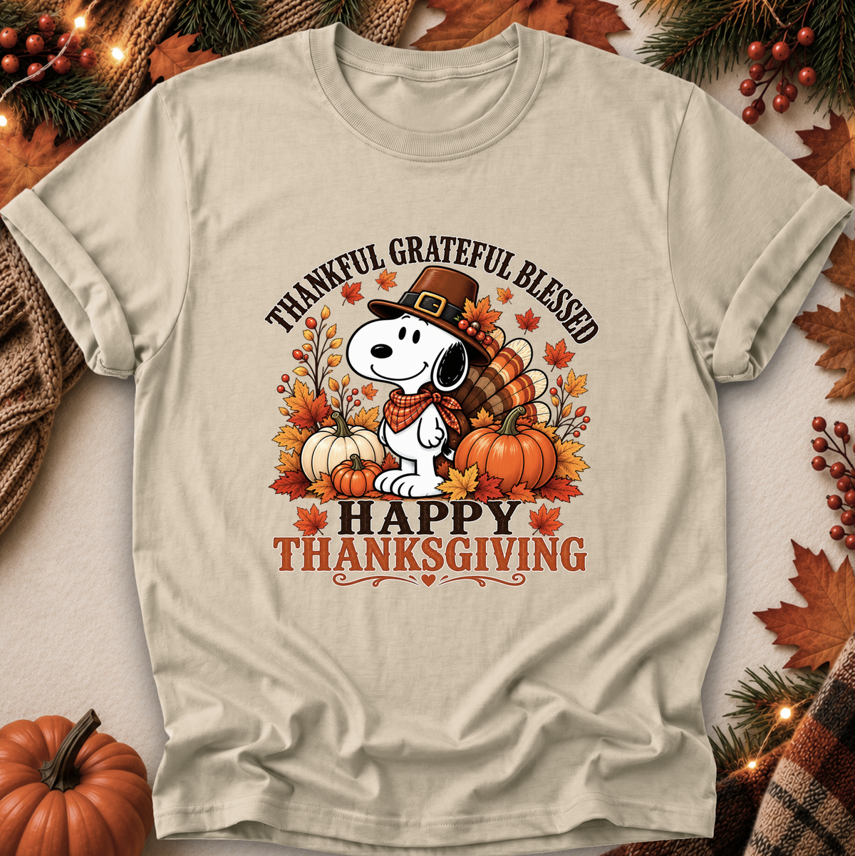 A beige graphic t-shirt featuring Snoopy and Woodstock in a Thanksgiving theme, surrounded by pumpkins, autumn leaves, and the text 'THANKFUL GRATEFUL BLESSED' and 'HAPPY THANKSGIVING'. The shirt is displayed in a flat-lay arrangement with seasonal props like pine branches, berries, and a small pumpkin.