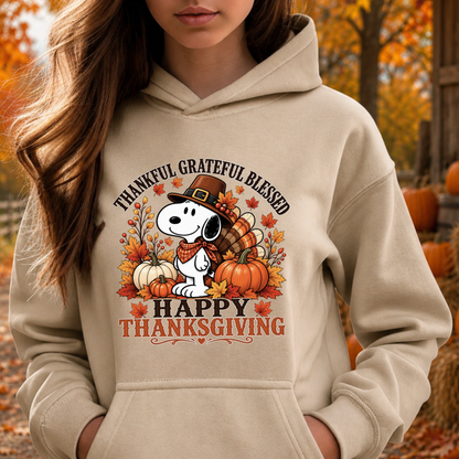A woman wearing a beige hoodie with a festive Thanksgiving graphic featuring Snoopy, pumpkins, autumn leaves, and the text 'THANKFUL GRATEFUL BLESSED HAPPY THANKSGIVING'. The background shows an outdoor autumn scene with trees and pumpkins.