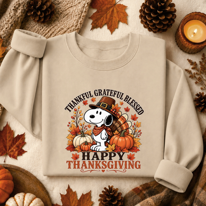 A beige long-sleeve crewneck sweatshirt featuring a festive Thanksgiving graphic with Snoopy, pumpkins, autumn leaves, and the text 'THANKFUL GRATEFUL BLESSED HAPPY THANKSGIVING'. The sweatshirt is styled in a cozy flat-lay arrangement with pinecones, fall leaves, a candle, and a knit blanket.