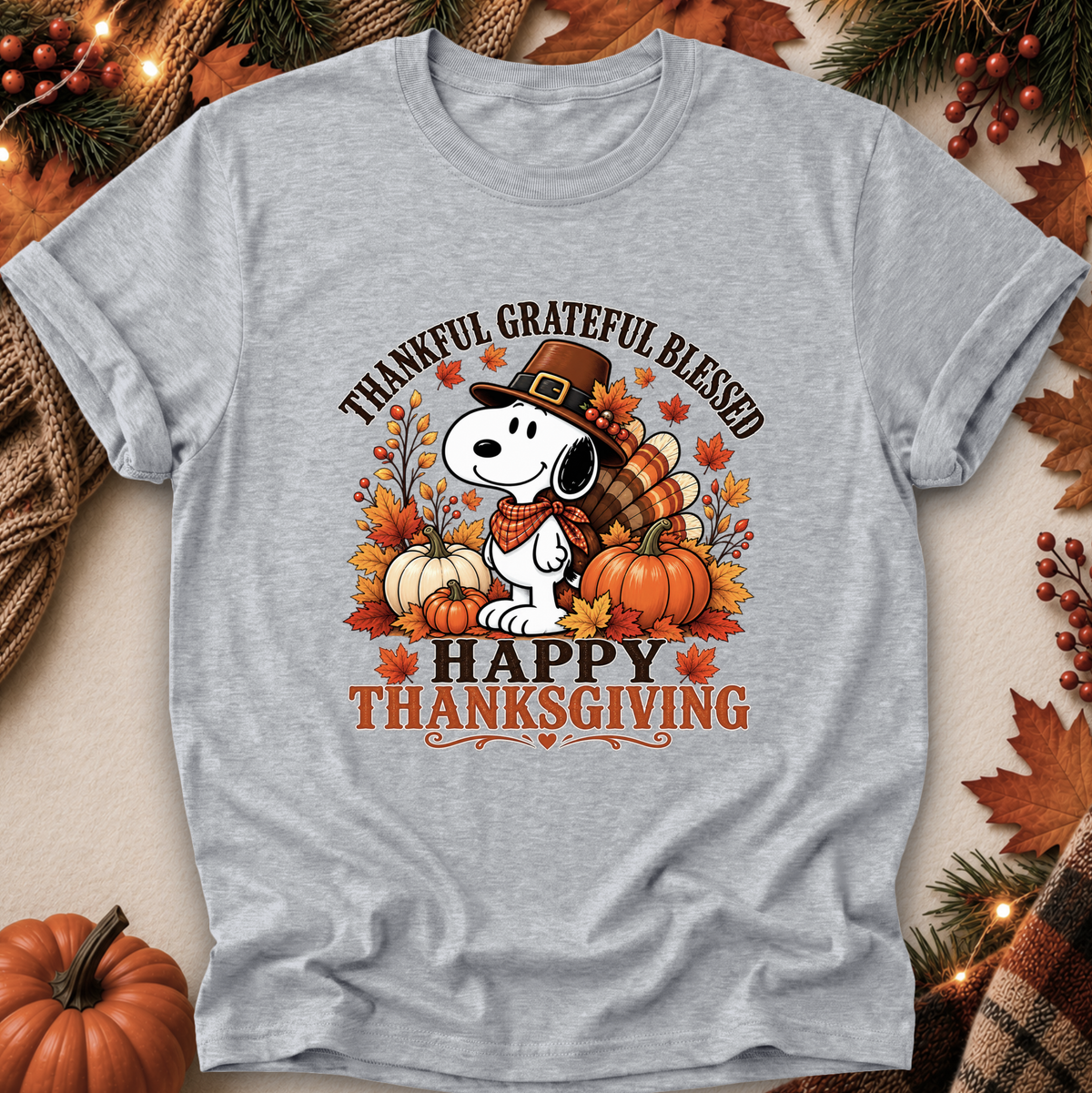 A heather gray short-sleeve t-shirt featuring a Thanksgiving-themed graphic of Snoopy wearing a pilgrim hat with pumpkins, autumn leaves, and the text 'THANKFUL GRATEFUL BLESSED HAPPY THANKSGIVING'. The shirt is displayed in a flat-lay arrangement with seasonal props like pine branches, berries, and a small pumpkin.