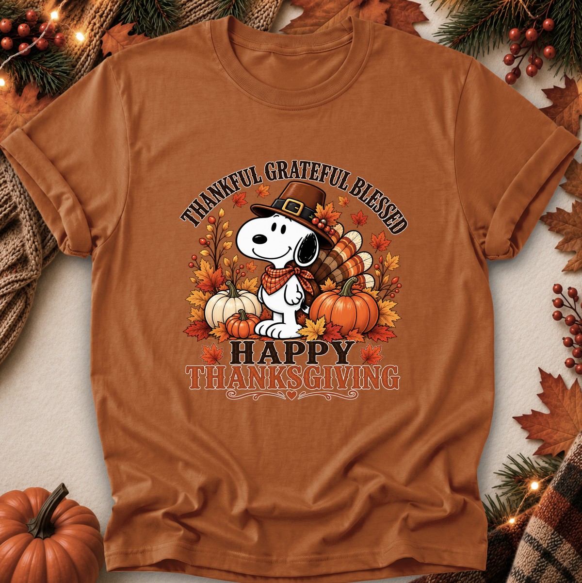 A rust-brown short-sleeve graphic T-shirt laid flat with seasonal props. The shirt features a Snoopy illustration wearing a pilgrim hat, surrounded by pumpkins, autumn leaves, and the text 'THANKFUL GRATEFUL BLESSED' and 'HAPPY THANKSGIVING.