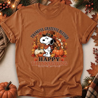 A rust-brown short-sleeve graphic T-shirt laid flat with seasonal props. The shirt features a Snoopy illustration wearing a pilgrim hat, surrounded by pumpkins, autumn leaves, and the text 'THANKFUL GRATEFUL BLESSED' and 'HAPPY THANKSGIVING.