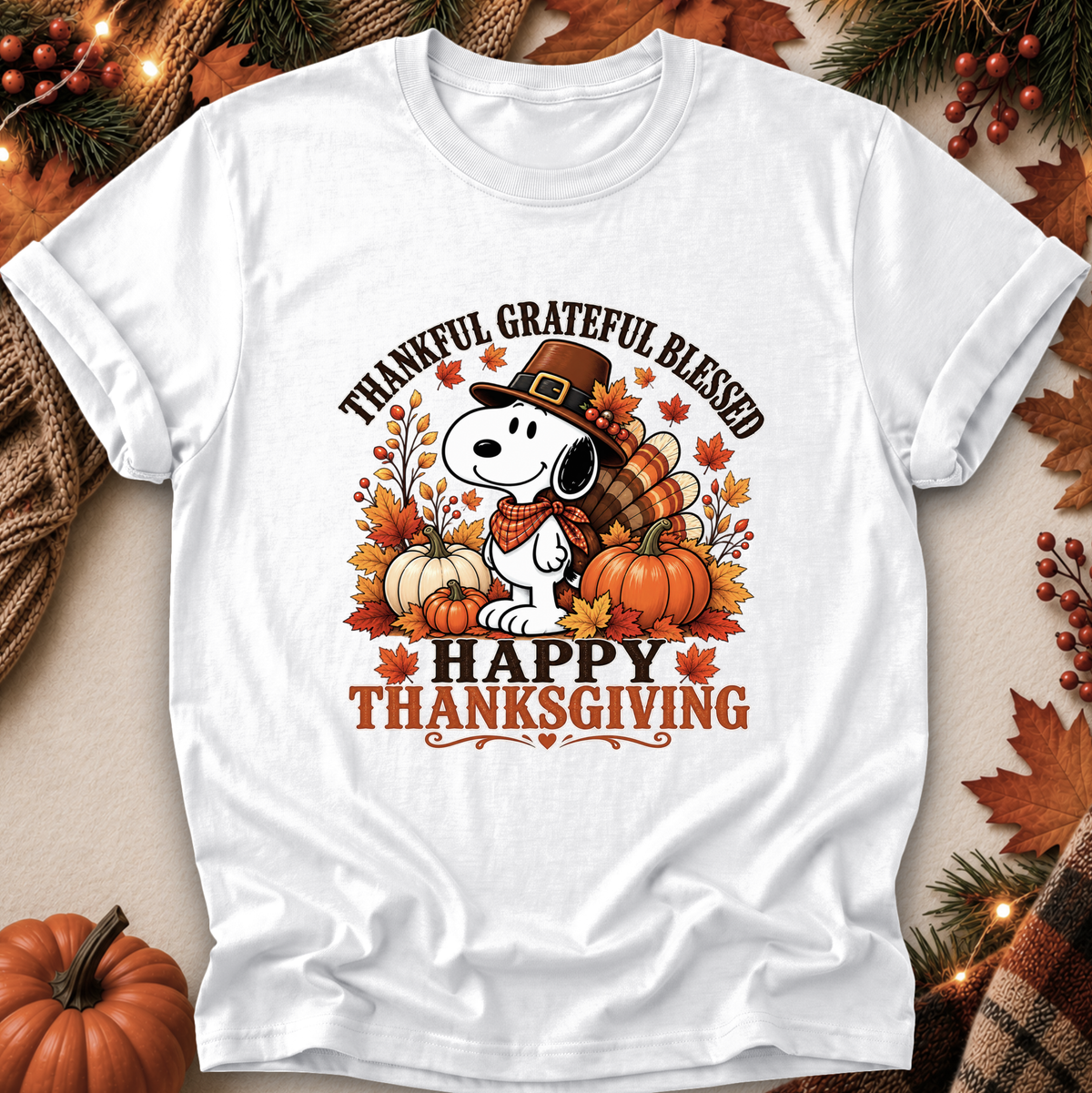 A white short-sleeve graphic T-shirt laid flat with autumn-themed props around it. The shirt features a cartoon dog wearing a pilgrim hat and scarf, surrounded by pumpkins, fall leaves, and the text 'THANKFUL GRATEFUL BLESSED' and 'HAPPY THANKSGIVING'.
