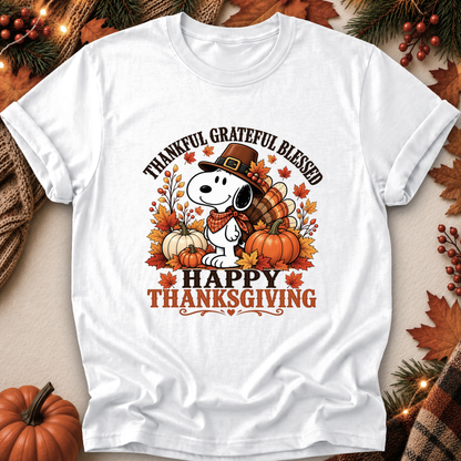 A white short-sleeve graphic T-shirt laid flat with autumn-themed props around it. The shirt features a cartoon dog wearing a pilgrim hat and scarf, surrounded by pumpkins, fall leaves, and the text 'THANKFUL GRATEFUL BLESSED' and 'HAPPY THANKSGIVING'.