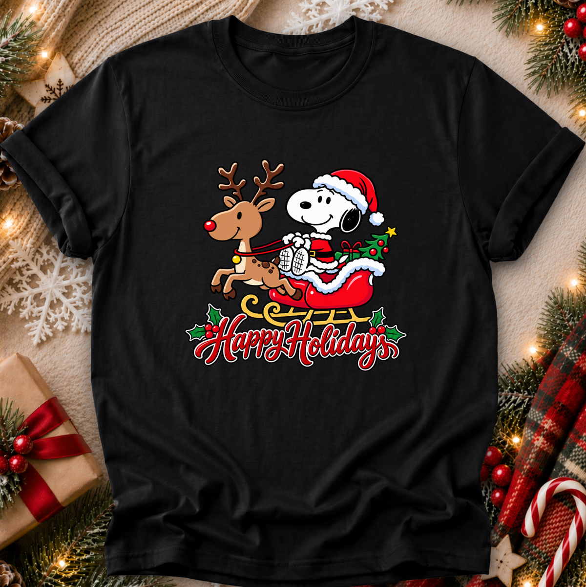 A black short-sleeve crewneck t-shirt featuring a festive holiday graphic of Snoopy in a Santa hat riding a sleigh with a reindeer and the text 'Happy Holidays' below. The shirt is displayed in a flat-lay setting surrounded by Christmas decorations including pine branches, lights, a wrapped gift, and a plaid blanket.