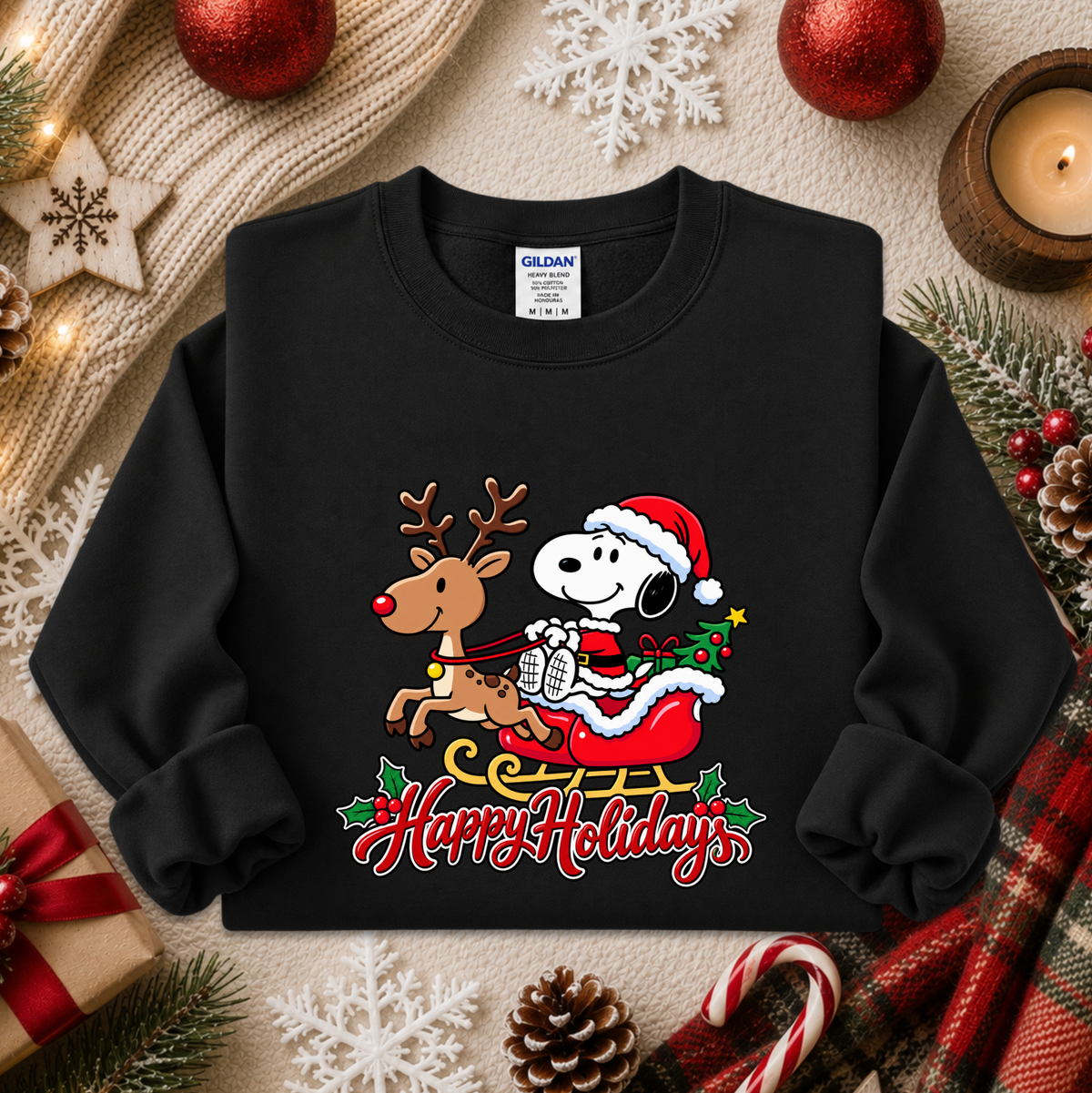 A black crewneck sweatshirt featuring a festive holiday graphic of Snoopy in a Santa hat riding a reindeer in a sleigh with 'Happy Holidays' written below. The sweatshirt is displayed in a styled flat-lay setting with Christmas decorations, pine branches, a candle, and a plaid scarf.