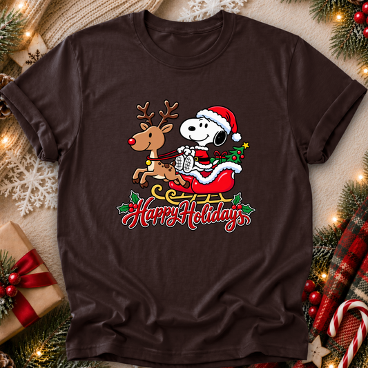 A dark brown short-sleeve crewneck t-shirt featuring a festive graphic of Snoopy in a Santa hat riding a sleigh with a reindeer and the words 'Happy Holidays' written below. The shirt is displayed in a flat-lay Christmas setting with pine branches, lights, a gift box, and a plaid blanket.