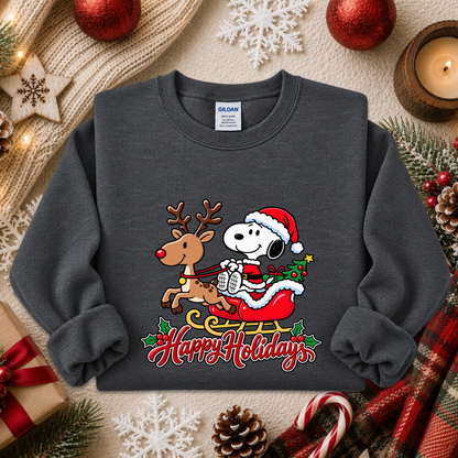 A dark gray crewneck sweatshirt featuring a festive graphic of Snoopy in a Santa hat riding a sleigh with a reindeer, with 'Happy Holidays' written below. The sweatshirt is styled in a cozy holiday flat-lay with red ornaments, pinecones, candy canes, and a knitted scarf.