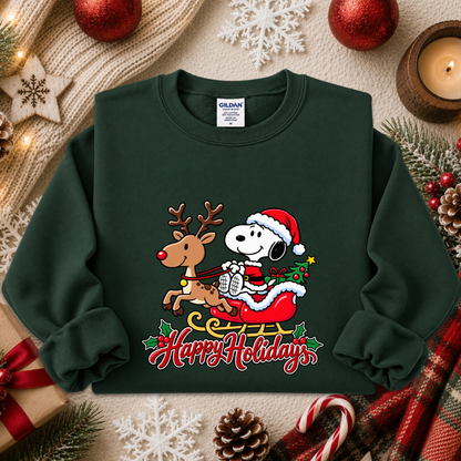 A dark green crewneck sweatshirt featuring a festive graphic of Snoopy in a Santa hat riding Santa Claus sleigh with a reindeer, under the text 'Happy Holidays'. The sweatshirt is styled in a cozy holiday flat-lay with a knit blanket, red ornaments, pinecones, and candy canes.