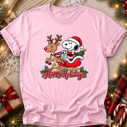 A light pink short-sleeve crewneck t-shirt featuring a festive graphic of Snoopy in a Santa hat riding a sleigh with a reindeer and the words 'Happy Holidays' written below. The shirt is displayed in a flat-lay holiday setting with pine branches, candy canes, a wrapped gift, and warm string lights.
