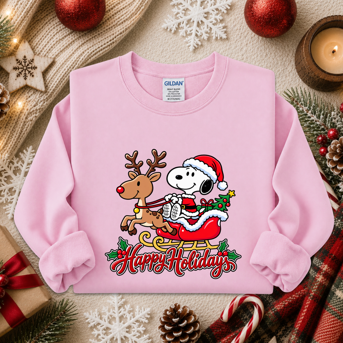A light pink long-sleeve crewneck sweatshirt featuring a festive graphic of Snoopy in a Santa hat and sleigh with a reindeer, under the words 'Happy Holidays'. The sweatshirt is styled in a cozy holiday flat-lay with a knitted scarf, red ornaments, a candle, and pinecones.