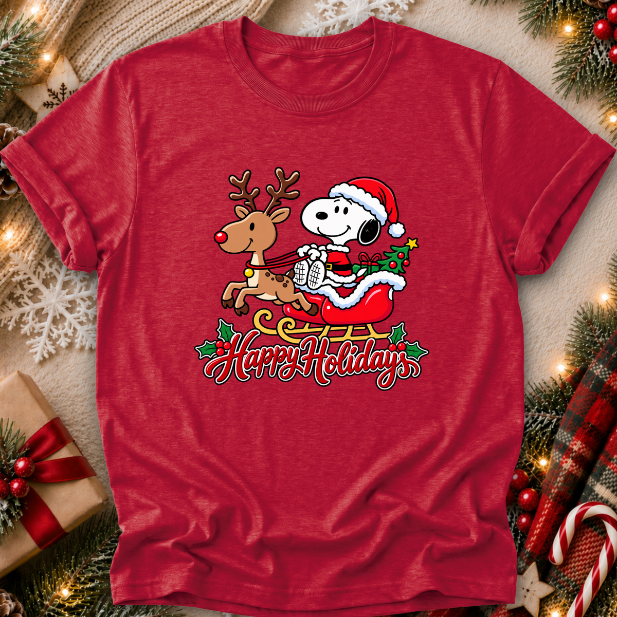 A red short-sleeve crewneck t-shirt featuring a festive graphic of Snoopy in a Santa hat riding a sleigh with a reindeer and the words 'Happy Holiday' written below. The shirt is displayed in a flat-lay setting decorated with Christmas lights, pine branches, a wrapped gift, and a plaid blanket.