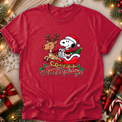 A red short-sleeve crewneck t-shirt featuring a festive graphic of Snoopy in a Santa hat riding a sleigh with a reindeer and the words 'Happy Holiday' written below. The shirt is displayed in a flat-lay setting decorated with Christmas lights, pine branches, a wrapped gift, and a plaid blanket.