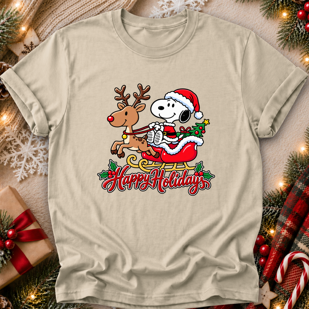 A beige graphic t-shirt featuring Snoopy and Woodstock in a Christmas sleigh with 'Happy Holidays' text, displayed in a cozy holiday flat-lay setting with pine branches, lights, and a gift box.