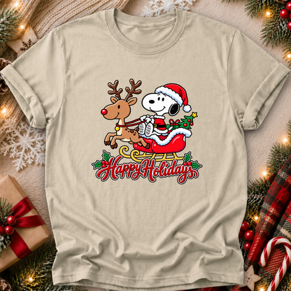A beige graphic t-shirt featuring Snoopy and Woodstock in a Christmas sleigh with 'Happy Holidays' text, displayed in a cozy holiday flat-lay setting with pine branches, lights, and a gift box.