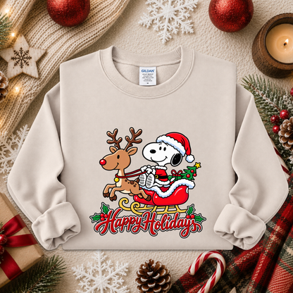 A light beige crewneck sweatshirt featuring a colorful graphic of Snoopy and Woodstock in a Santa-decorated sleigh with the words 'Happy Holidays' below, styled in a cozy holiday flat-lay with red ornaments, pinecones, a candle, and a knitted scarf.