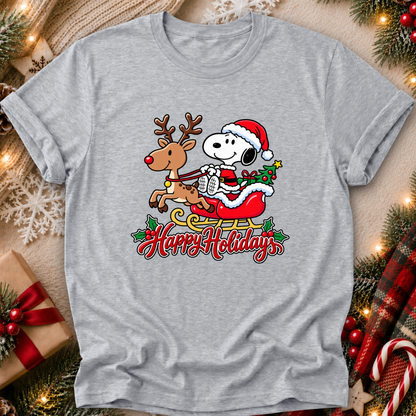 A heather gray short-sleeve t-shirt featuring a festive graphic of Snoopy in a Santa hat riding a sleigh with a reindeer, with 'Happy Holidays' written below. The shirt is styled in a cozy holiday flat-lay setting with pine branches, a wrapped gift, and warm string lights.
