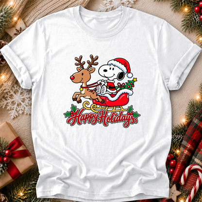 A white short-sleeve crewneck t-shirt featuring a festive graphic of Snoopy in a Santa hat riding a sleigh with a reindeer and the words 'Happy Holidays' written below. The shirt is displayed in a flat-lay setting surrounded by Christmas decorations including pine branches, a wrapped gift box, and a plaid blanket.