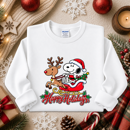 A white long-sleeve crewneck sweatshirt featuring a colorful graphic of Snoopy in a Santa hat riding a reindeer on a sleigh with 'Happy Holidays' written below. The sweatshirt is displayed in a styled flat-lay setting with holiday decorations including pine branches, red ornaments, a candle, and a gift box.