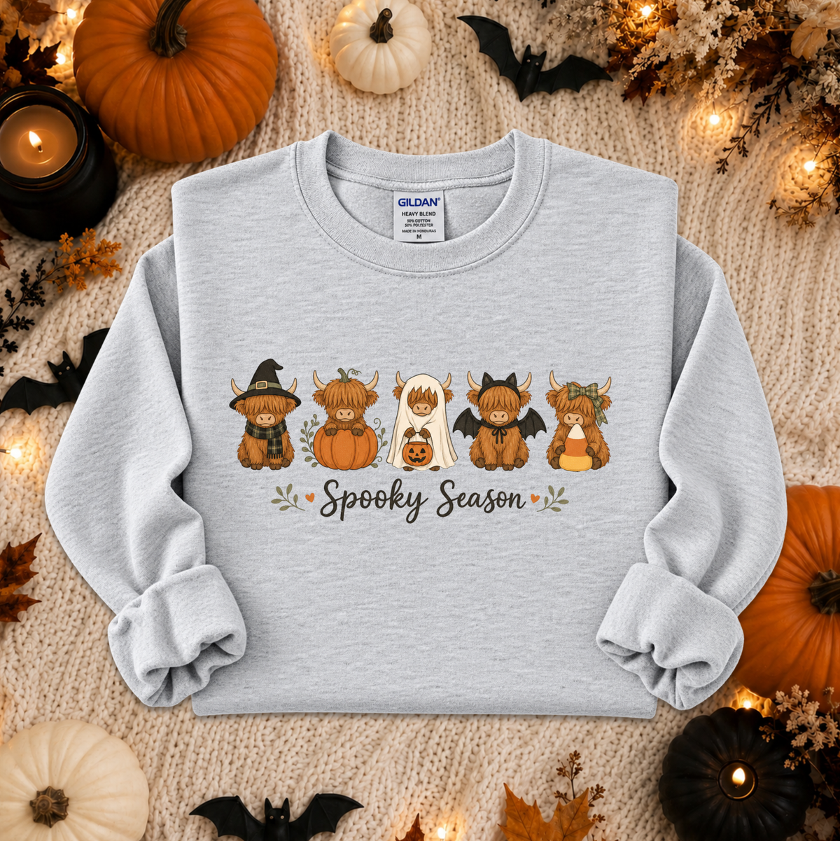 A light gray sweatshirt laid flat on a cozy beige knit blanket, surrounded by Halloween decorations including pumpkins, candles, leaves, and bats. The sweatshirt has a graphic of five cute cartoon pumpkins in autumn costumes with the words 'Spooky Season' underneath.