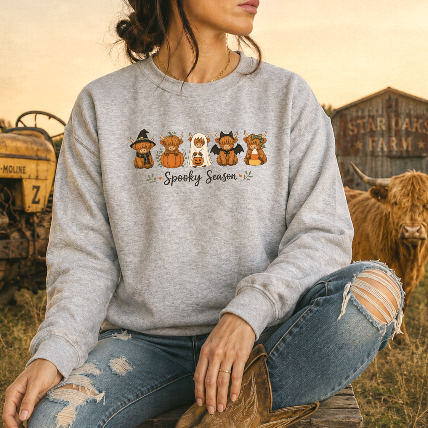 A woman wearing a heather gray crewneck sweatshirt with a 'Spooky Season' highland cow cartoon graphic, paired with ripped blue jeans and cowboy boots, sitting in a rustic outdoor setting with a cow and barn in the background.