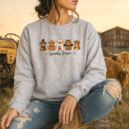 A woman wearing a heather gray crewneck sweatshirt with a 'Spooky Season' highland cow cartoon graphic, paired with ripped blue jeans and cowboy boots, sitting in a rustic outdoor setting with a cow and barn in the background.