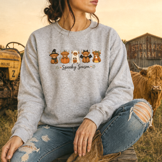 A woman wearing a heather gray crewneck sweatshirt with a 'Spooky Season' highland cow cartoon graphic, paired with ripped blue jeans and cowboy boots, sitting in a rustic outdoor setting with a cow and barn in the background.