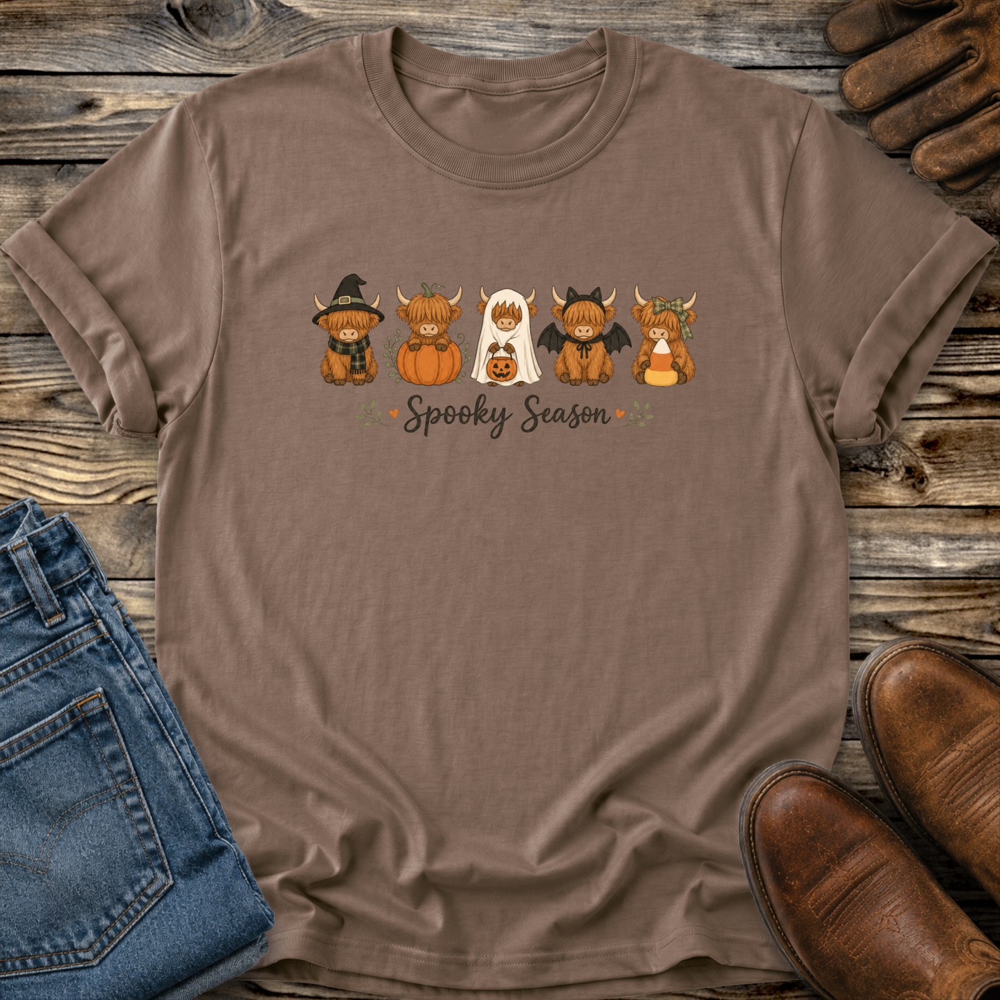 A brown short-sleeve crewneck t-shirt laid flat on a rustic wooden surface, featuring a 'Spooky Season' graphic with five cute cartoon highland cows in Halloween costumes. The shirt is styled with blue jeans, brown leather shoes, and a pair of work gloves.