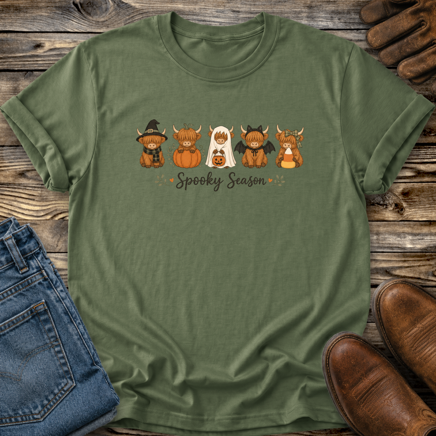 An olive green short-sleeve t-shirt with a 'Spooky Season' highland cows cartoon graphic, styled in a flat-lay on a rustic wooden background with jeans, brown shoes, and leather gloves.