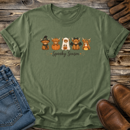 An olive green short-sleeve t-shirt with a 'Spooky Season' highland cows cartoon graphic, styled in a flat-lay on a rustic wooden background with jeans, brown shoes, and leather gloves.