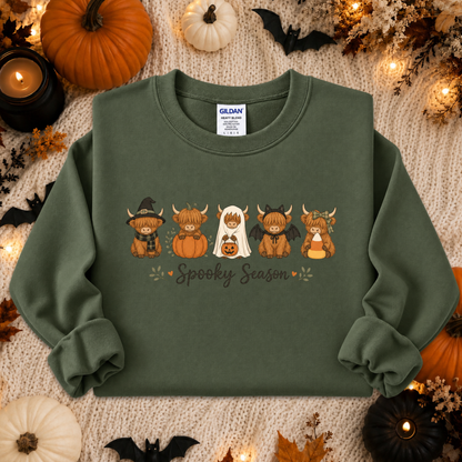 An olive green crewneck sweatshirt with a 'Spooky Season' cartoon graphic, laid flat on a cozy white knit blanket surrounded by Halloween decorations including pumpkins, candles, and bats.