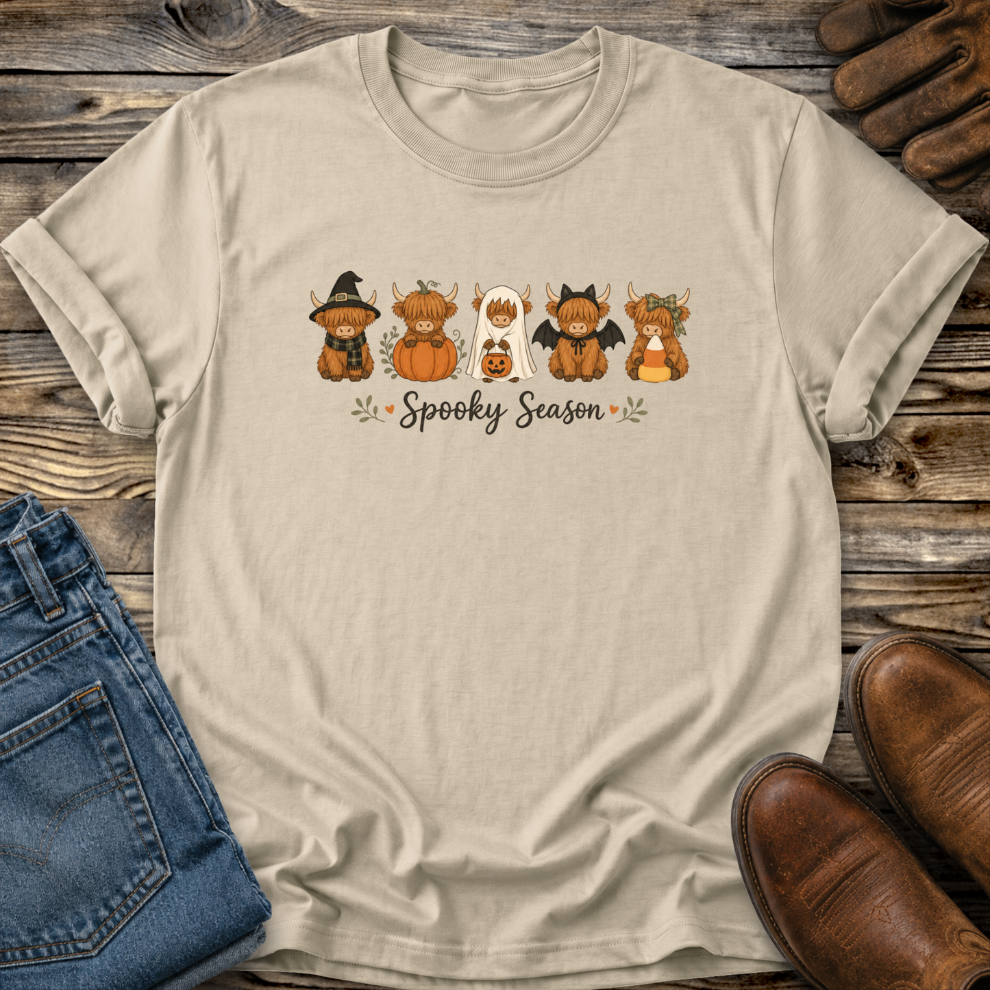A beige graphic t-shirt laid flat on a rustic wooden surface, styled with blue jeans, brown shoes, and leather gloves. The t-shirt features a cartoon graphic of five cute highland cow Halloween characters and the text 'Spooky Season'.