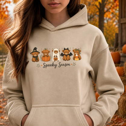 A woman wearing a beige hoodie with a 'Spooky Season' cartoon graphic of five Scottish highlands in Halloween costumes, set against an autumn outdoor background with pumpkins and orange foliage.