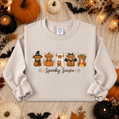 A light beige crewneck sweatshirt featuring a 'Spooky Season' graphic with cartoon Scottish terriers in Halloween costumes, styled in a flat lay with pumpkins, candles, and autumn leaves.
