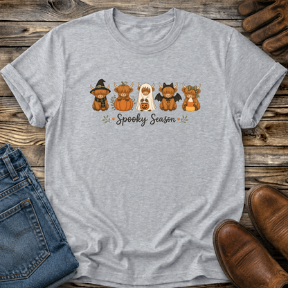 A heather gray short-sleeve T-shirt laid flat on a rustic wood surface, featuring a Halloween-themed graphic with five cartoon highland cow characters in costumes and the words 'Spooky Season.' Blue jeans, brown shoes, and a pair of wooden gloves are partially visible around it as styling props.