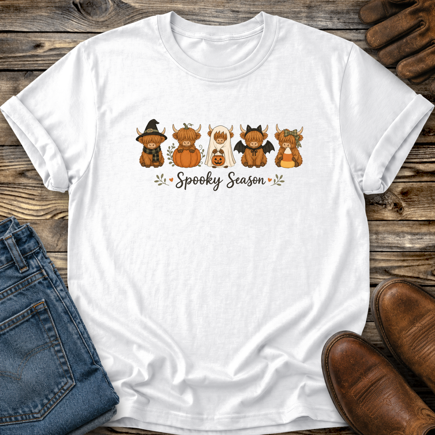 A white short-sleeve T-shirt laid flat on a rustic wood surface, featuring a colorful graphic of five cute cartoon highland cows dressed in Halloween costumes with the words 'Spooky Season' underneath. Blue jeans, brown shoes, and a pair of gloves are partially visible around the shirt as styling props.