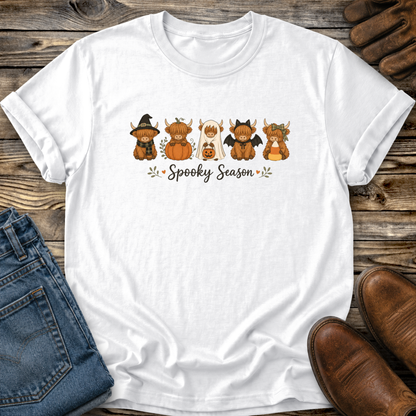 A white short-sleeve T-shirt laid flat on a rustic wood surface, featuring a colorful graphic of five cute cartoon highland cows dressed in Halloween costumes with the words 'Spooky Season' underneath. Blue jeans, brown shoes, and a pair of gloves are partially visible around the shirt as styling props.