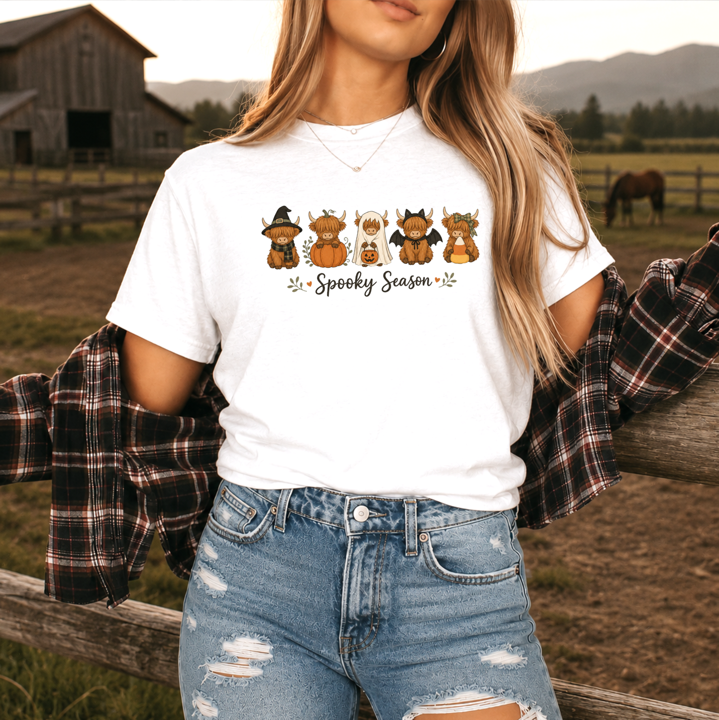 A woman wearing a white graphic t-shirt with a 'Spooky Season' highland cow cartoon design, paired with distressed denim shorts and a plaid flannel shirt draped off her shoulders, set against a rustic barn and field background.
