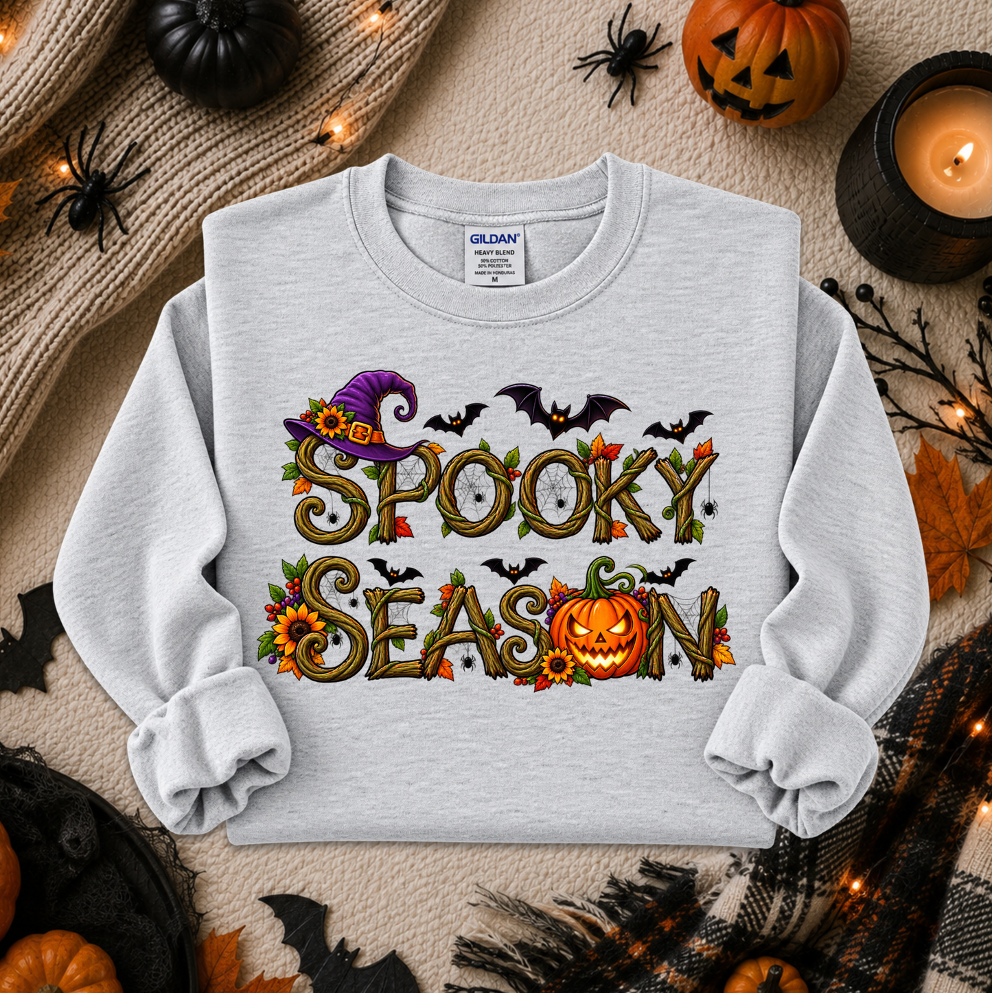 A light gray crewneck sweatshirt with a colorful 'Spooky Season' graphic, laid flat on a cozy beige blanket surrounded by Halloween props including pumpkins, spiders, a candle, and a knit sweater.