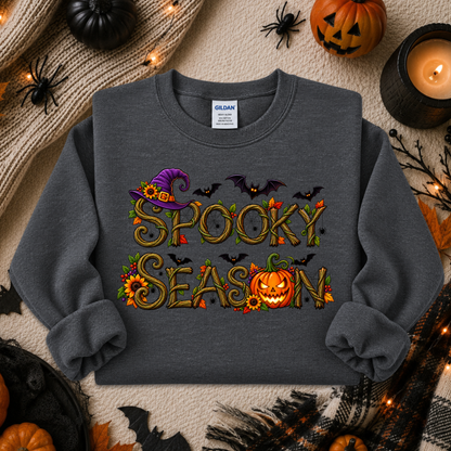 A dark gray crewneck sweatshirt with a 'Spooky Season' graphic, styled in a flat lay with Halloween props including pumpkins, a candle, and spider decorations. The brand Gildan is visible on the inner neck label.