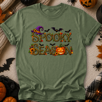 An olive green short-sleeve t-shirt with a 'Spooky Season' graphic print, laid flat on a textured beige blanket surrounded by Halloween props like pumpkins, a black spider, and a witch hat.