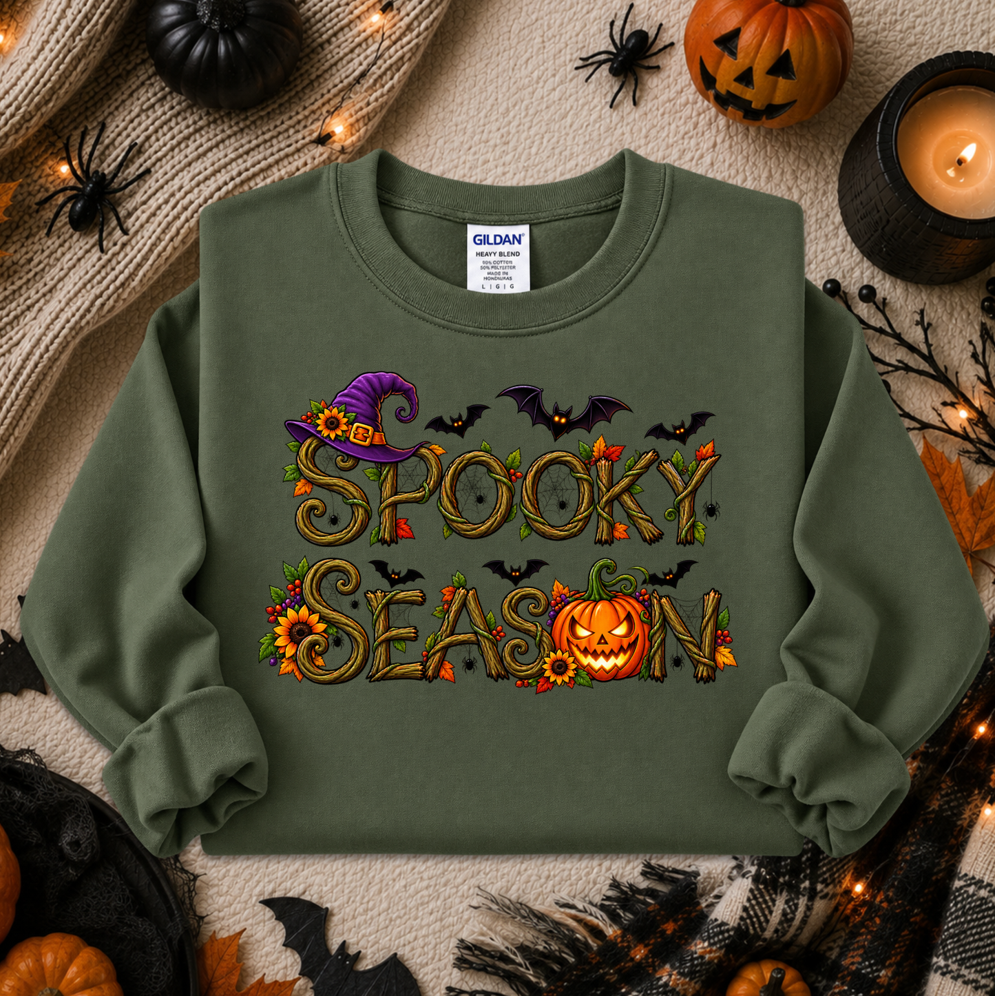 An olive green crewneck sweatshirt with a 'Spooky Season' graphic, styled in a flat lay with Halloween props including pumpkins, a candle, and spider decorations. The brand Gildan is visible on the inner neck label.