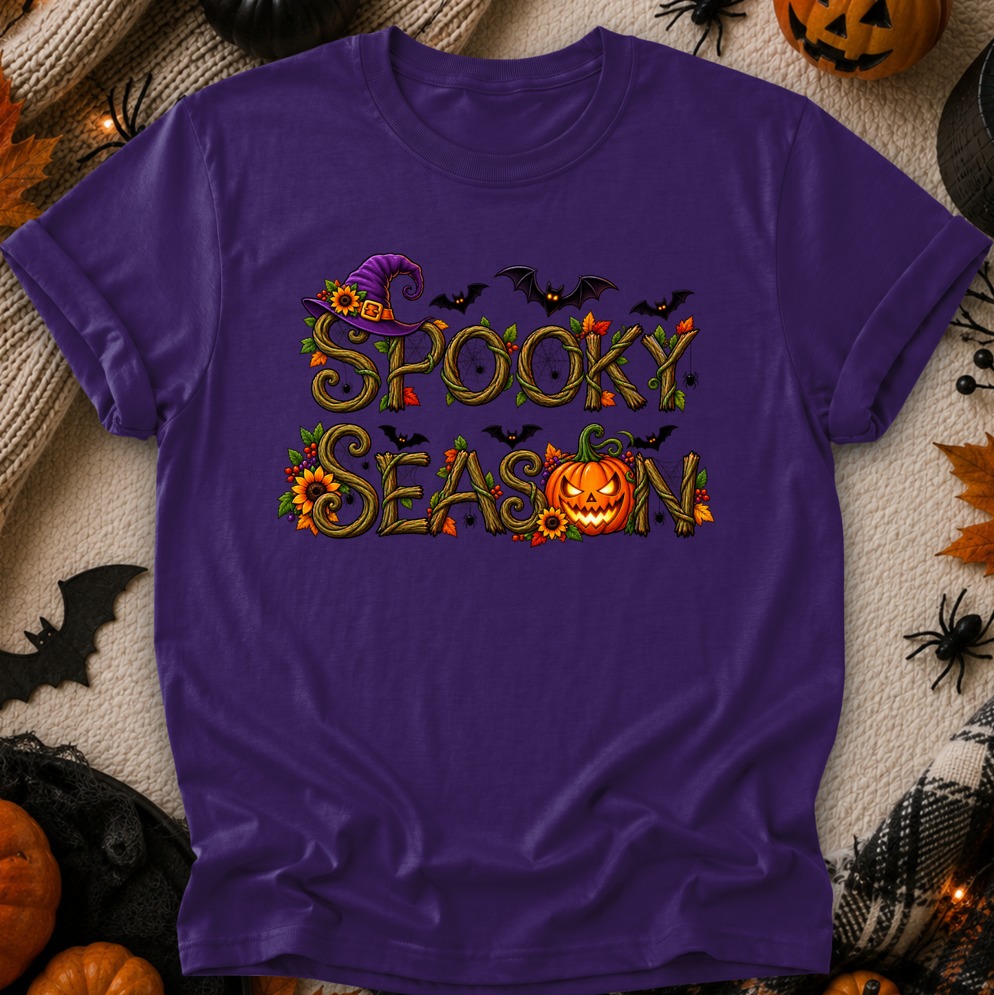 A purple short-sleeve t-shirt with a 'Spooky Season' graphic, laid flat on a cozy beige blanket surrounded by Halloween decorations like pumpkins, spiderwebs, and black plastic bats.