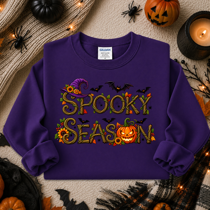 A purple long-sleeve crewneck sweatshirt with a 'Spooky Season' graphic, laid flat on a beige knit blanket surrounded by Halloween decorations including pumpkins, spiders, and a candle.