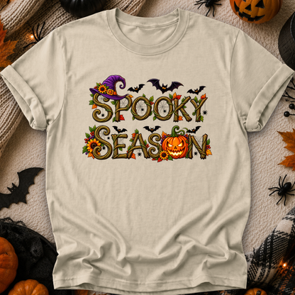 An off-white t-shirt with a 'Spooky Season' graphic print, laid flat on a cozy autumn-themed background with pumpkins, spiderwebs, and a plaid blanket.