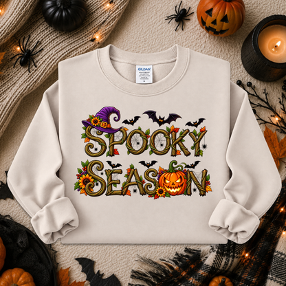 A cream-colored crewneck sweatshirt featuring a 'Spooky Season' graphic with pumpkins, bats, and a witch hat, laid flat on a cozy autumn-themed background with pumpkins, a candle, and a knit blanket.
