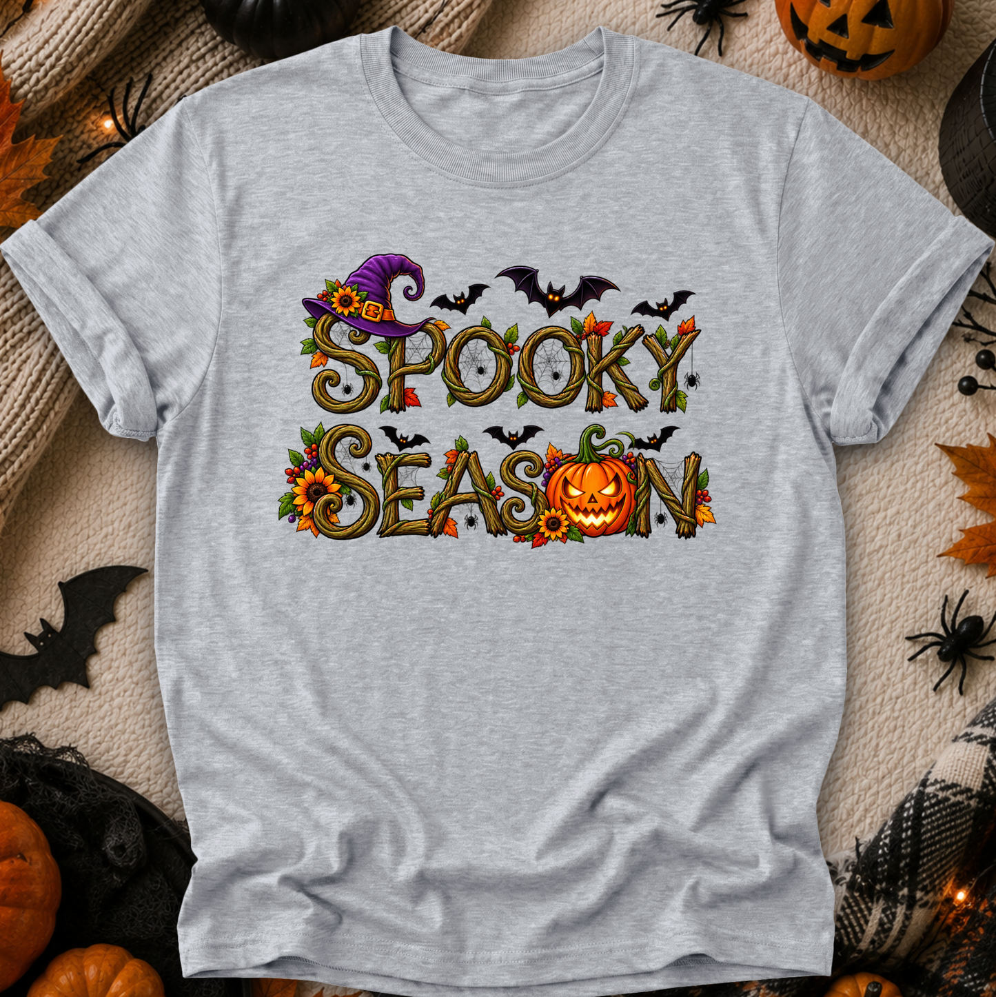 A light heather gray short-sleeve t-shirt with a 'Spooky Season' graphic print, laid flat on a textured beige blanket surrounded by Halloween props like pumpkins, a spider, and a witch hat.