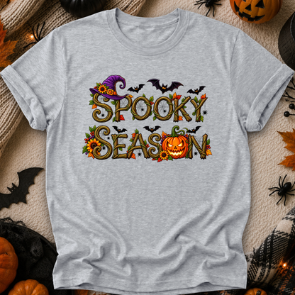 A light heather gray short-sleeve t-shirt with a 'Spooky Season' graphic print, laid flat on a textured beige blanket surrounded by Halloween props like pumpkins, a spider, and a witch hat.