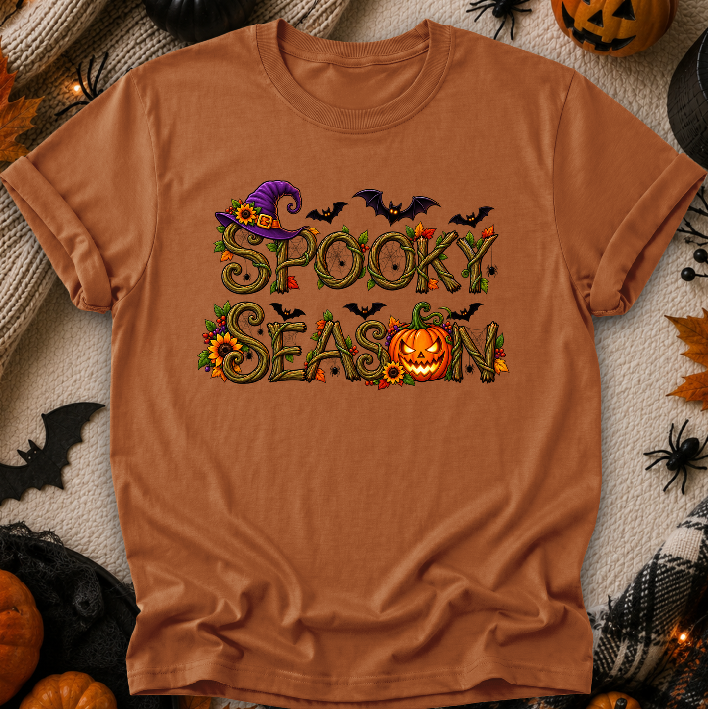 A rust-orange crewneck t-shirt featuring a 'Spooky Season' graphic with a witch hat, bats, sunflowers, and a jack-o'-lantern, laid flat on a cozy beige knit blanket surrounded by Halloween props.